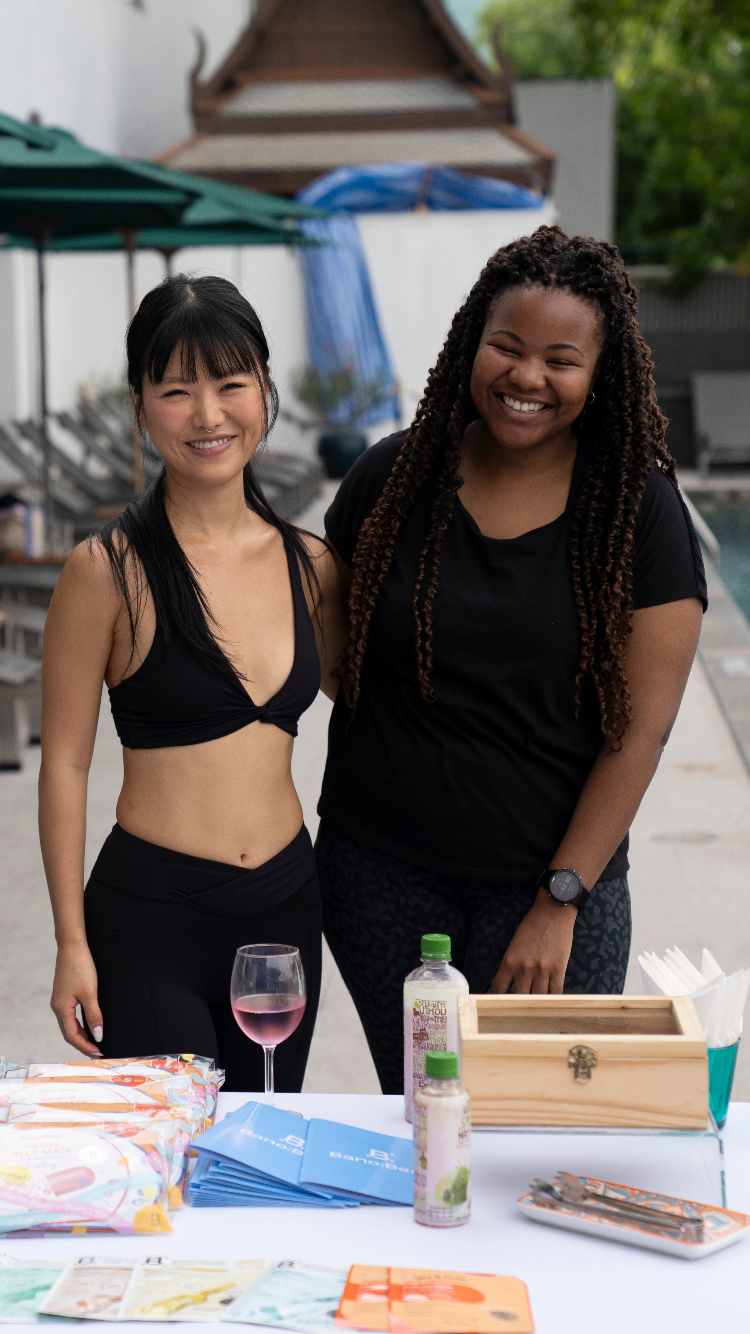 Jean and Rejoice standing behind a table with products and a drink, outdoor setting.